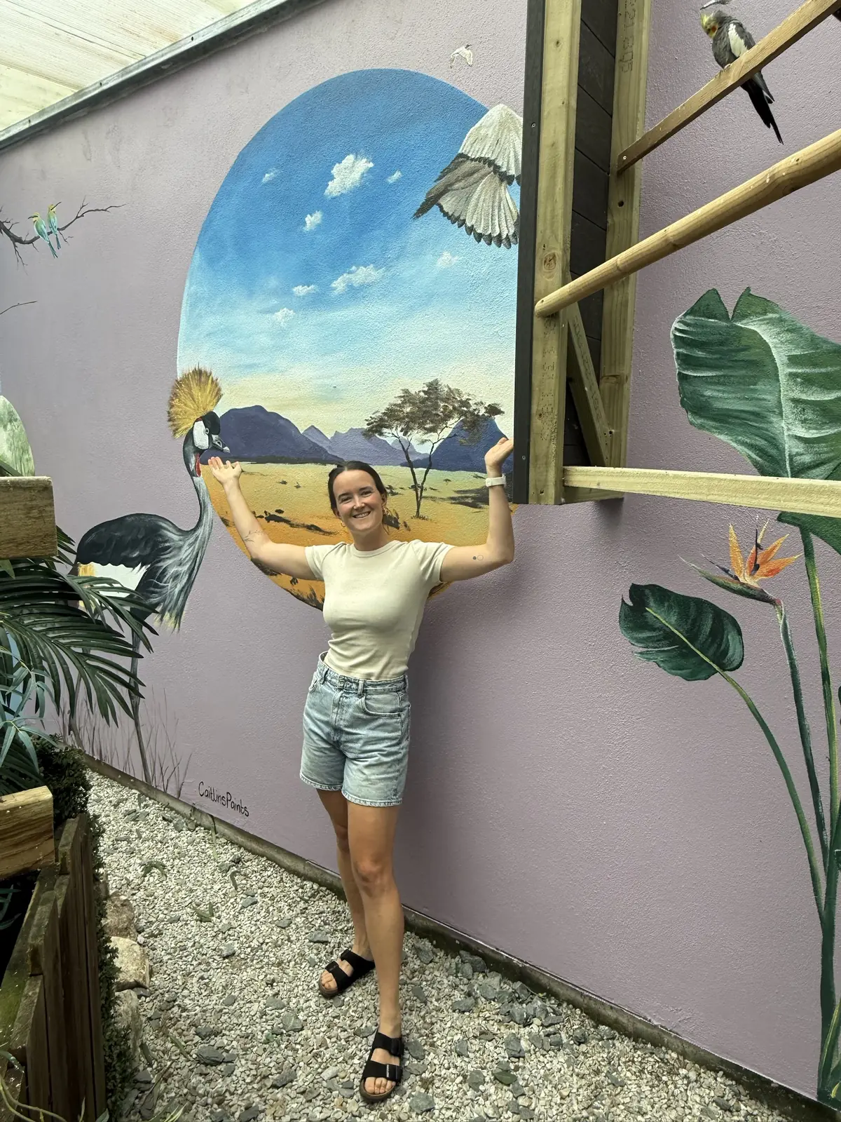 Caitlin standing with her arms raised in front of her painted crowned crane and savanna mural